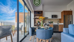 Een moderne keuken met een magnetron, koelkast en houten kasten. Er is een eethoek met een glazen tafel en blauwe stoelen.