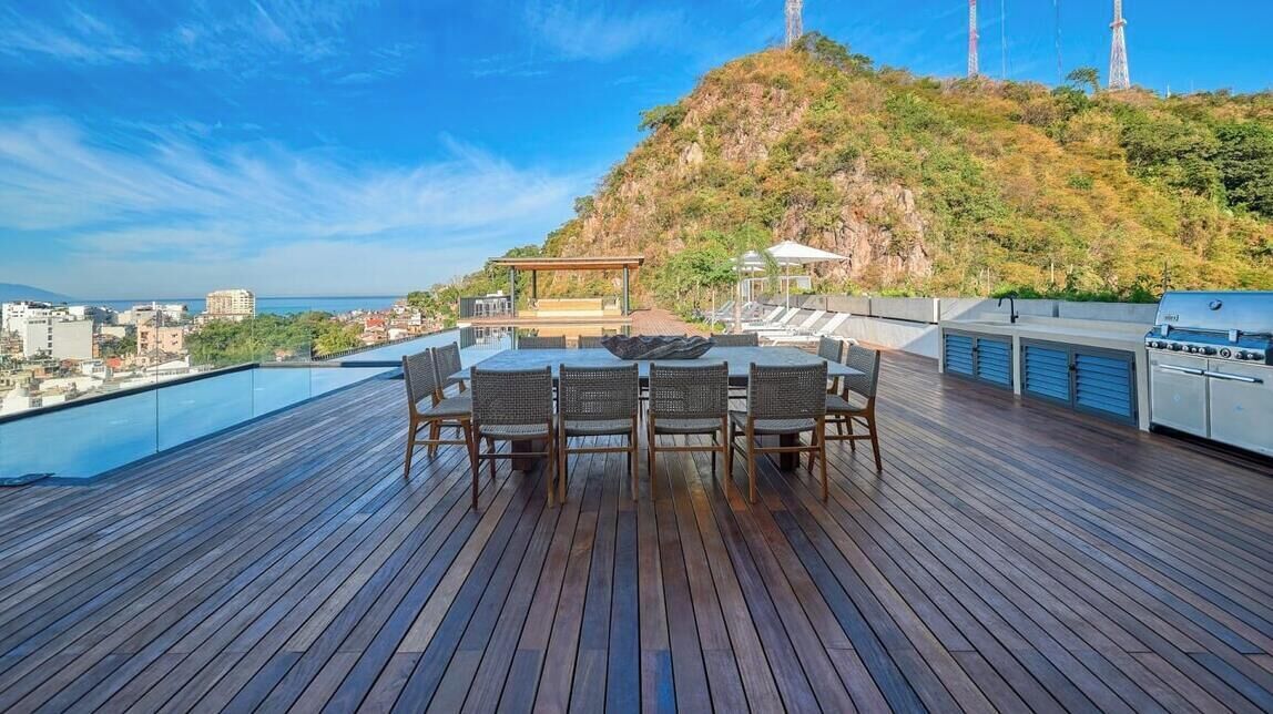 A rooftop terrace with a dining table and chairs, a grill, and a view of a cityscape and hills.