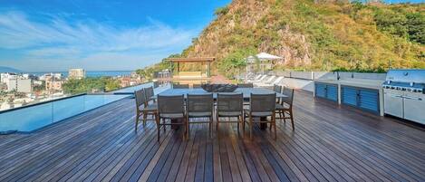 A rooftop terrace with a dining table and chairs, a grill, and a view of a cityscape and hills.