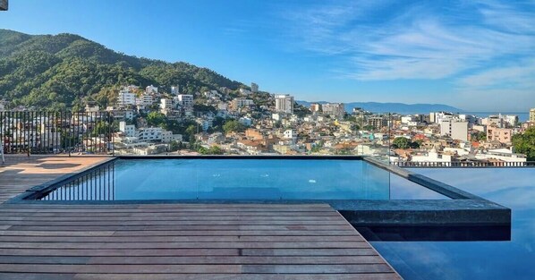 Une piscine à débordement située sur le toit, avec une terrasse en bois, offrant une vue panoramique sur la ville, ses bâtiments et une montagne.