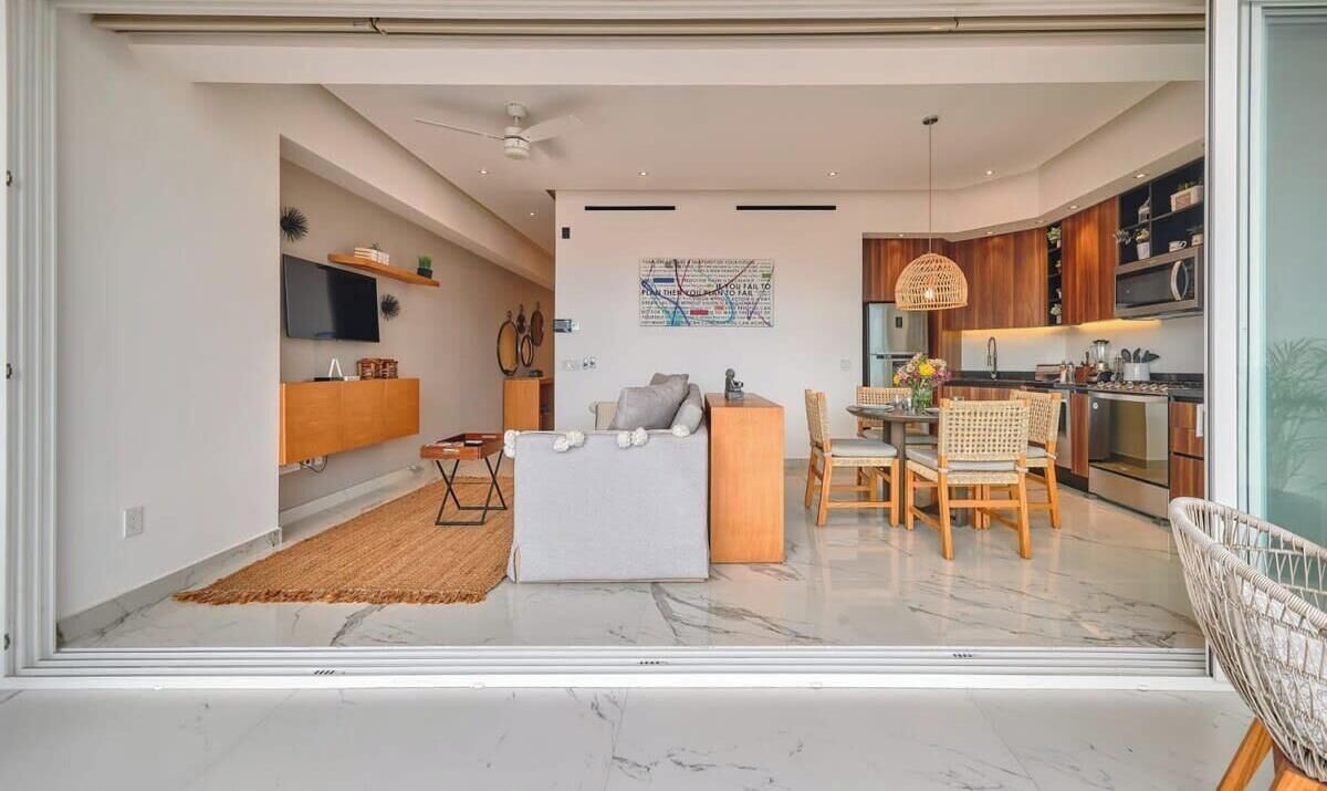 Una sala de estar moderna con un sofá, una mesa de comedor con sillas, una zona de cocina con armarios y electrodomésticos, y un ventanal grande.
