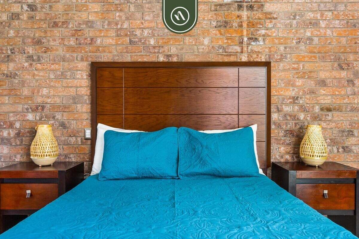 Ein Hotelzimmer mit Backsteinwand, Holz-Kopfteil, blauer Bettdecke und zwei Nachttischen mit dekorativen Lampen.