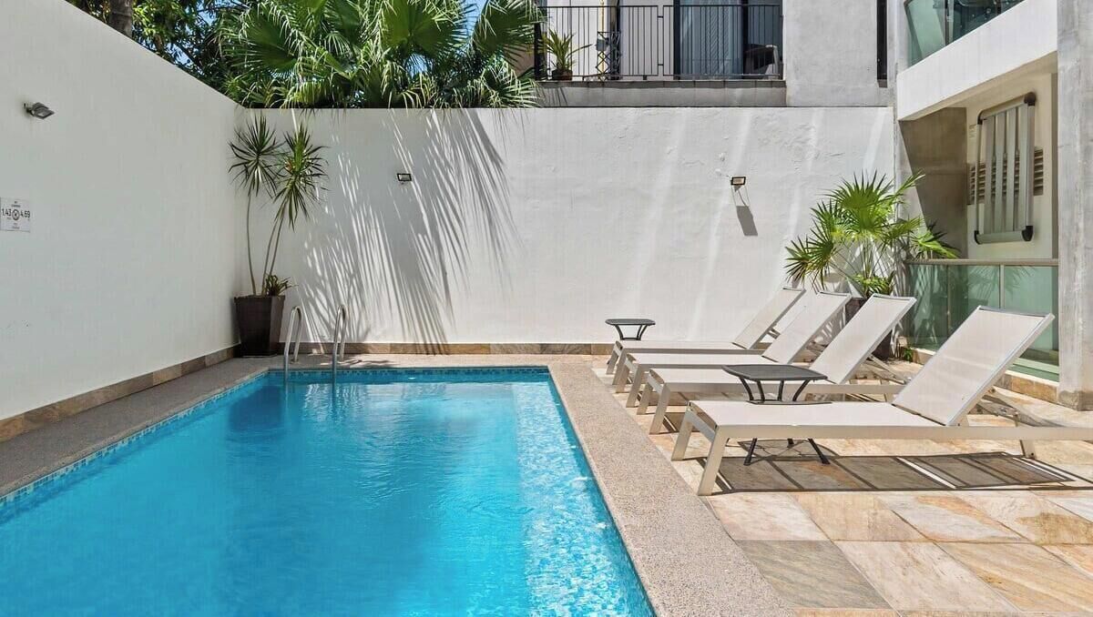 A swimming pool with lounge chairs, potted plants, and a white wall.