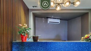 Una zona de recepción con un mostrador azul con textura, un panel de madera vertical, una planta en maceta, una cesta pequeña y una unidad de aire acondicionado instalada en el techo.