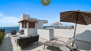 A rooftop terrace with a view of the sea, a small building with a tiled roof, and outdoor furniture including a sofa and a table. - Brand New 1 BR Condo Right Next to the Malecon (Puerto Vallarta)