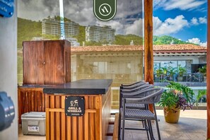 A restaurant with a wooden counter, wicker chairs, and a view of buildings and greenery. - Luxury 1 BR Condo in Romantic Zone - Infinity Pool (Puerto Vallarta)