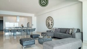Una sala de estar moderna con un sofá gris, otomanas y una mesa de centro. Se observa un reloj de pared grande.