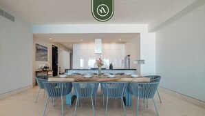 Un comedor moderno con una mesa de madera y sillas azules, una cocina al fondo y un ventanal grande.