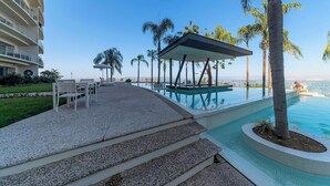 Zona de piscina con escalones que llevan a un área de descanso, palmeras y un edificio con balcones.