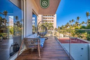 A balcony with a wicker sofa, a hanging chair, and a view of palm trees and a building.