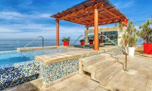 Uma área de piscina com pérgola, escadas que dão acesso à piscina e plantas em vasos.