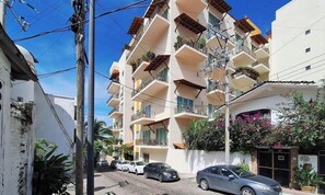 Um edifício residencial com vários andares e varandas, rua pavimentada, carros estacionados e céu azul claro.