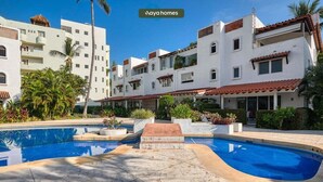 A pool area with a clear blue pool, white buildings with red-tiled roofs, and lush greenery.