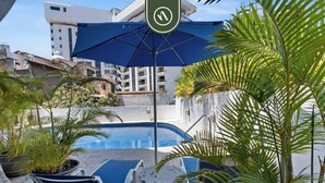 A swimming pool area with lounge chairs and umbrellas, surrounded by potted plants and modern buildings in the background. - Stunning 2 BR Condo - Oceanview Terrace - Downtown (Puerto Vallarta)