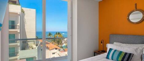 A hotel room with a large sliding glass door leading to a balcony with a view of the ocean and buildings.