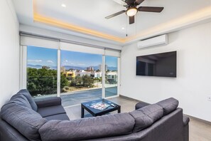 A modern living room with a large window, a ceiling fan, and a flat-screen TV. - Believe Condo Flamingos Believe 302 (Bucerías)