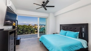 A modern bedroom with a bed, a ceiling fan, a TV, and a balcony with a city view. - Believe Condo Flamingos Believe 302 (Bucerías)