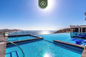 An infinity pool with clear blue water, a view of the ocean, and a building with columns in the background. - Amazing House With Private Terrace - Private Pool (Cabo San Lucas)