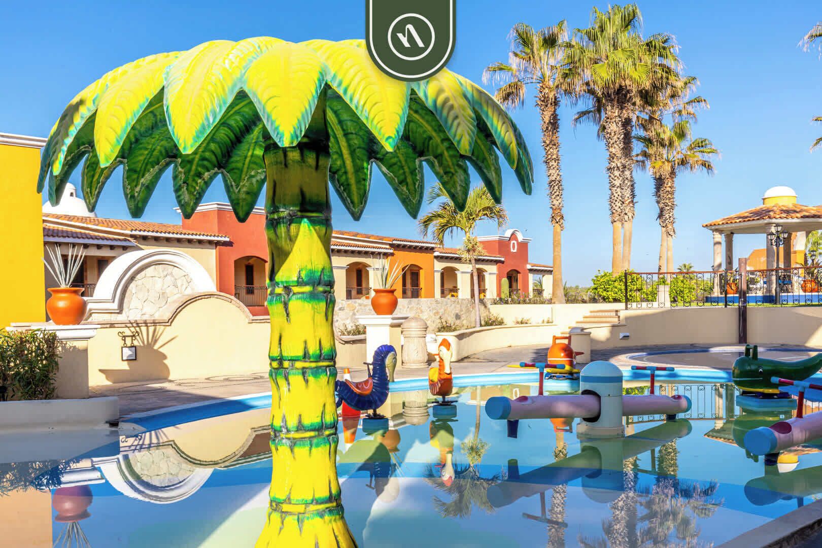 A pool area with a large palm tree sculpture, a clear blue sky, and a building with a tiled roof in the background.