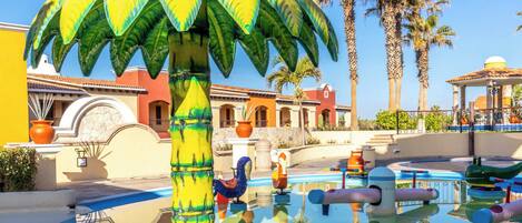 A pool area with a large palm tree sculpture, a clear blue sky, and a building with a tiled roof in the background.