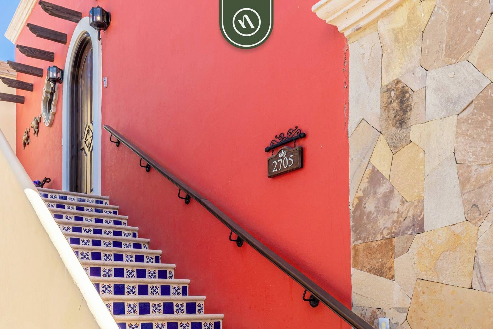 Un bâtiment rouge avec un escalier menant à une porte, une enseigne portant le numéro 2705, et une enseigne circulaire verte avec un « V » blanc au centre.