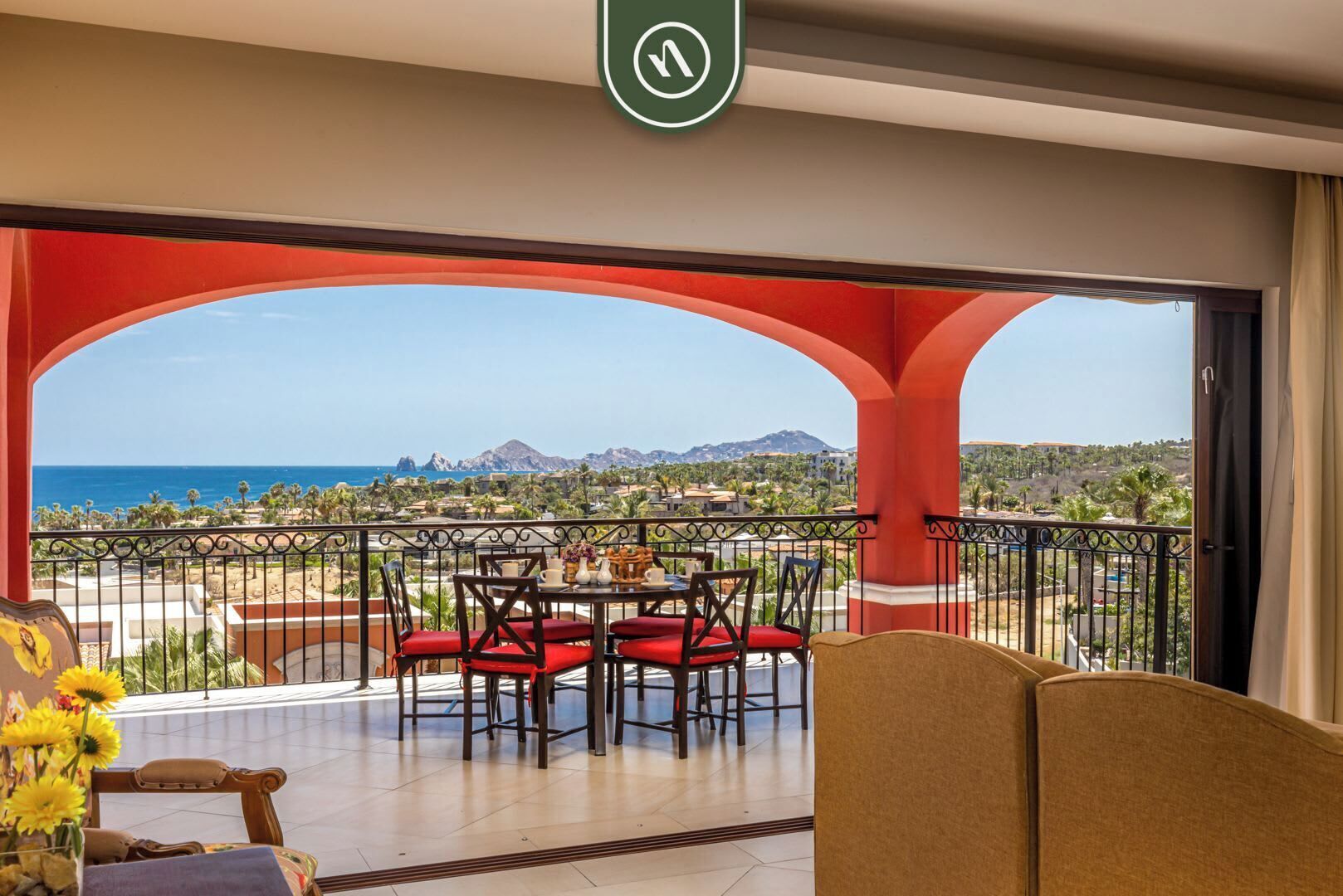 Un balcón con mesa y sillas, vistas al mar y montañas, y un arco rojo.