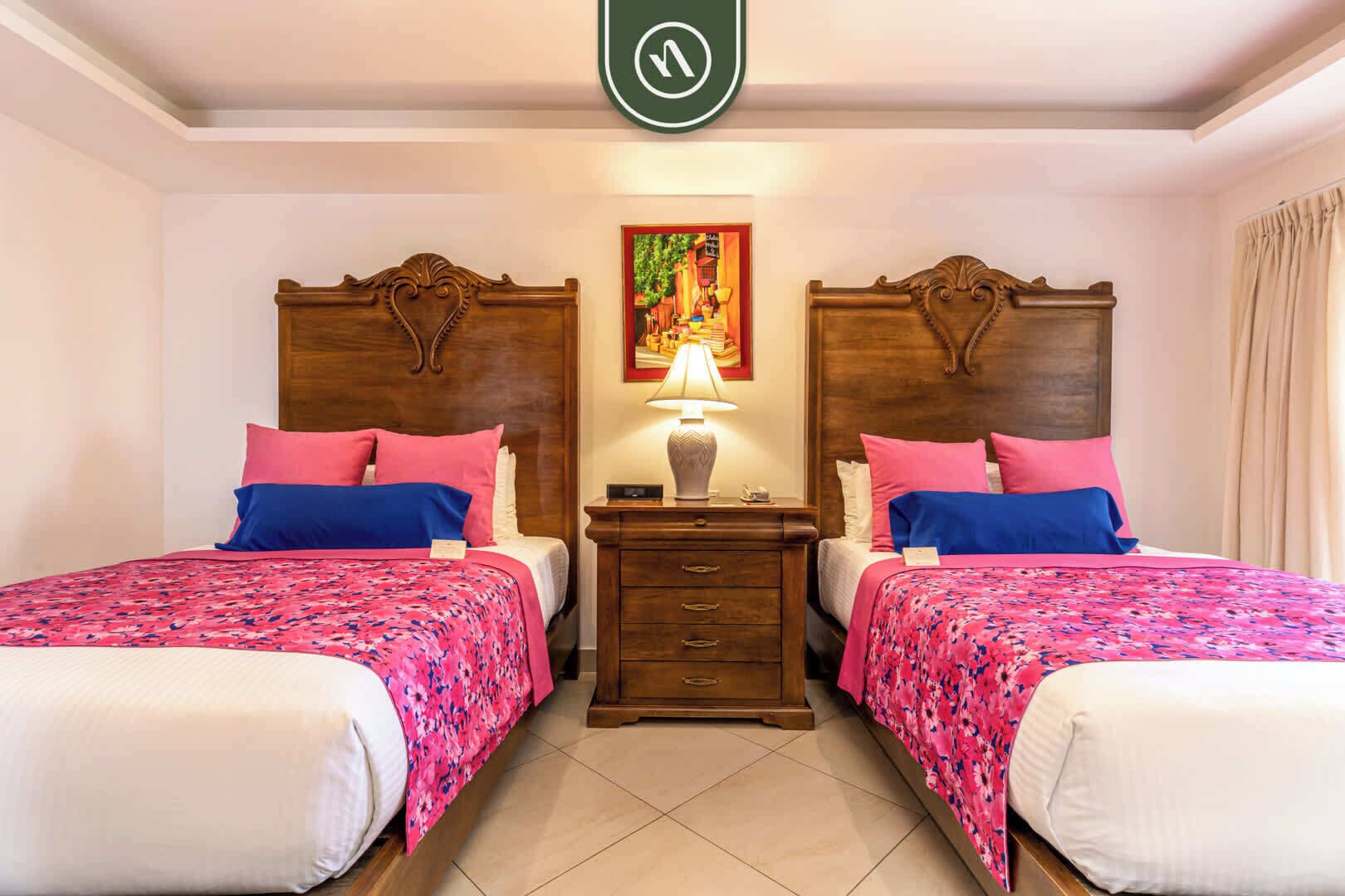 A hotel room with two beds, each with a floral bedspread and matching pillows. There is a nightstand with a lamp between the beds.
