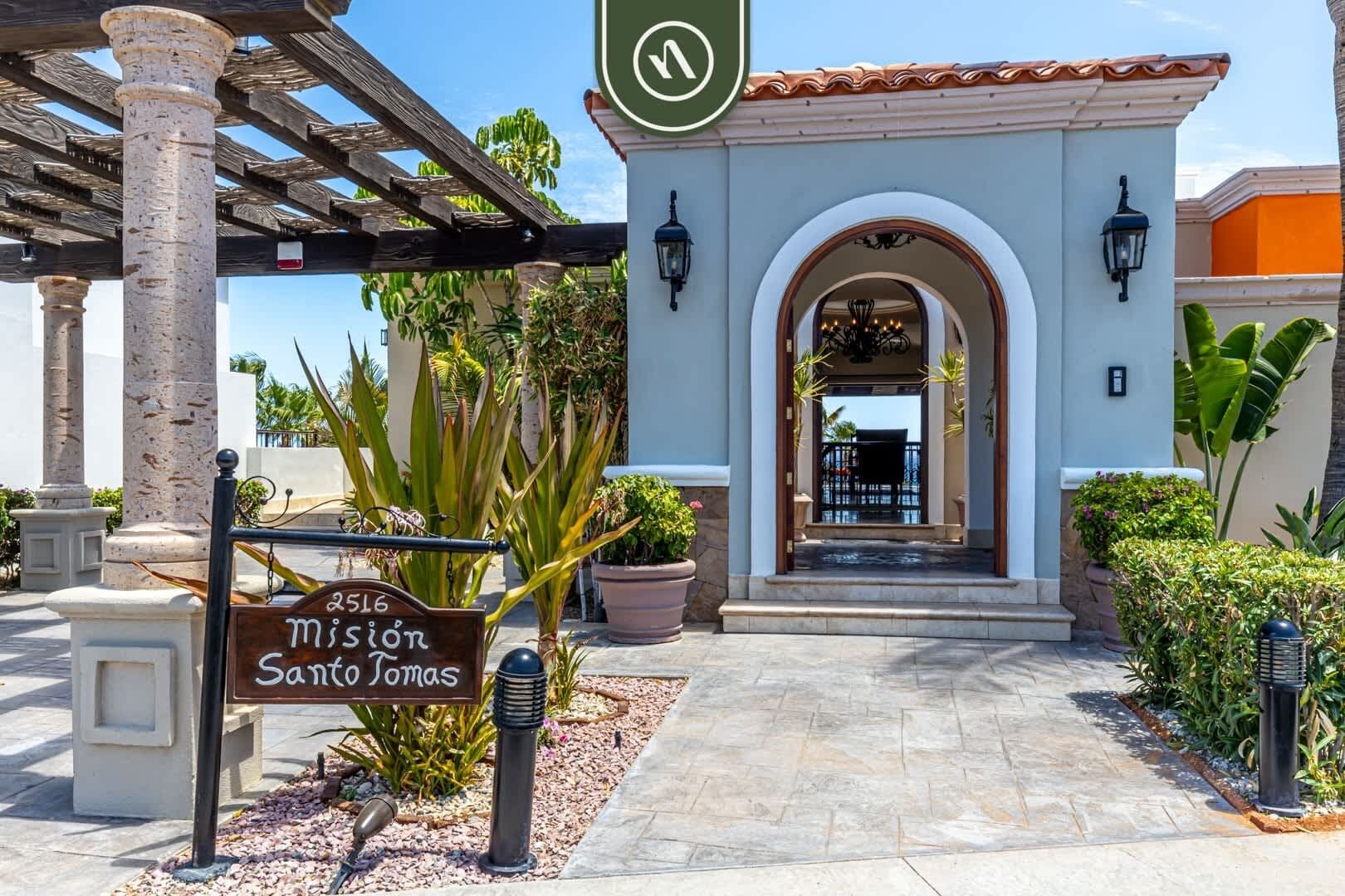 A hotel entrance with a sign reading "Mission Santa Tomas," a covered walkway, and a view of the interior through an arched doorway.