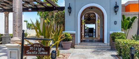 A hotel entrance with a sign reading "Mission Santa Tomas," a covered walkway, and a view of the interior through an arched doorway.