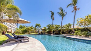 A clear swimming pool with lounge chairs and umbrellas, surrounded by palm trees and a building in the background. - New 2 BR Condo in San Jose - Pool - Tennis (San José del Cabo)