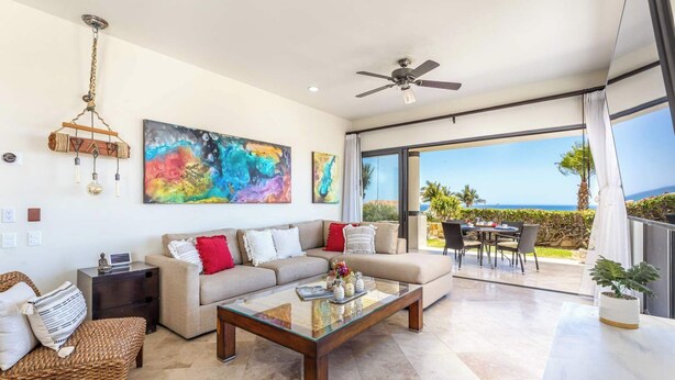 A modern living room with a sofa, coffee table, and artwork, leading to a balcony with a view of the sea. - New 2 BR Condo in San Jose - Pool - Tennis (San José del Cabo)