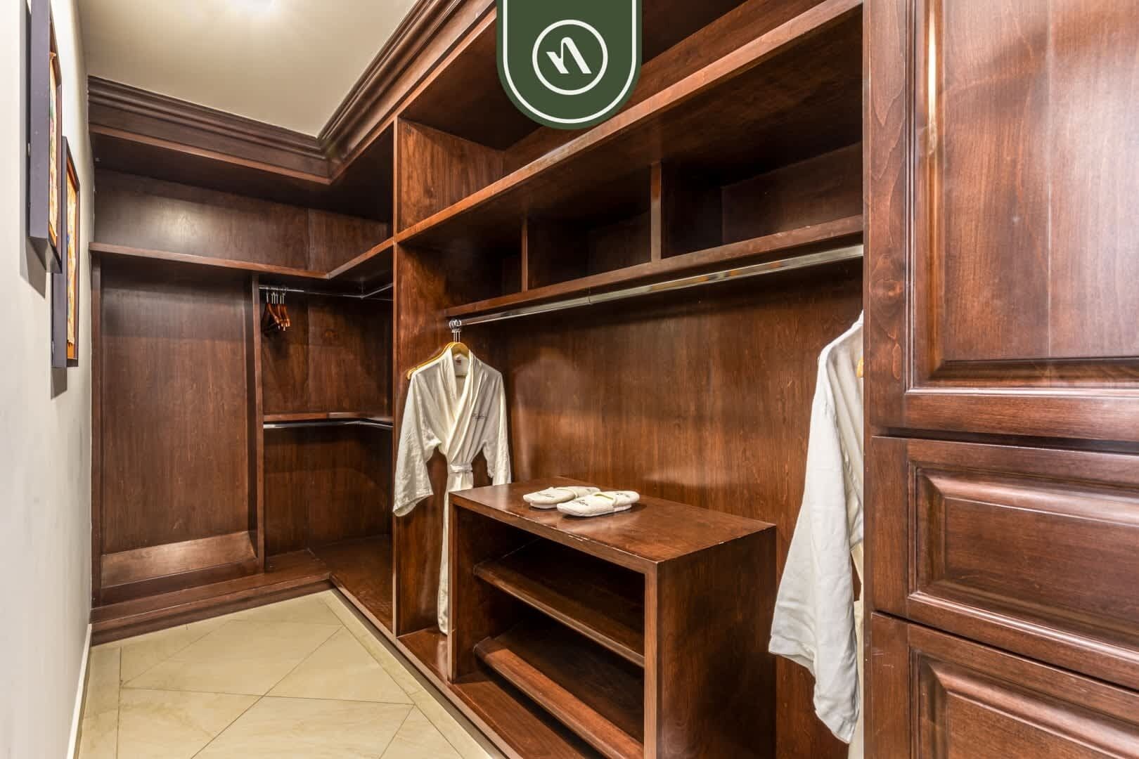 Un vestidor de madera con estantes y una barra para colgar ropa, una bata blanca colgada en la barra, y una mesita con un objeto blanco encima.