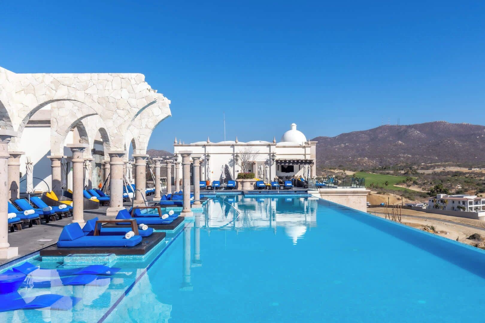 Una piscina infinita con sillas reclinables azules, un edificio blanco con cúpula y montañas al fondo.