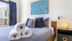 A hotel room with a bed, pillows, a bedside table with a lamp, a wall-mounted painting, and a window with curtains. - Amazing 2BR Condo With Rooftop Pool - BBQ (Cabo San Lucas)