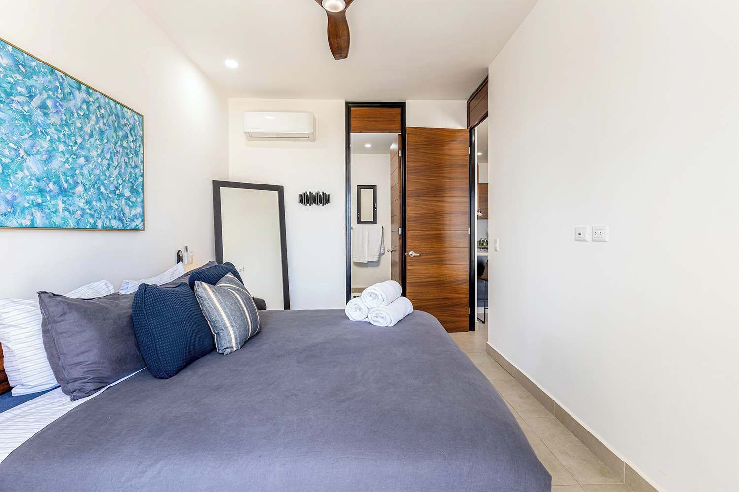 A modern bedroom with a large bed, a ceiling fan, an air conditioning unit, a wooden door, and a bathroom visible through an open door.