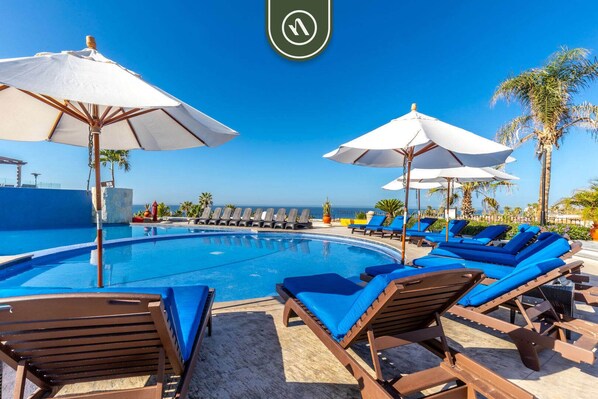 A poolside area with lounge chairs and umbrellas, a clear view of the ocean, and a stone wall. - Great House in Cabo With Ocean View - Pool (Cabo San Lucas)