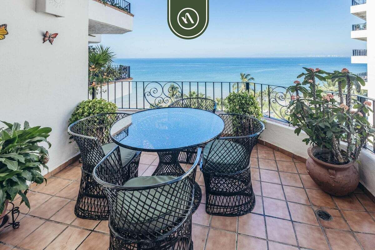 A balcony with a round table and chairs, a potted plant, and a view of the ocean.