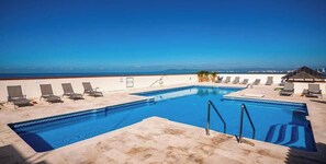 A rectangular pool with a clear blue waterline, surrounded by lounge chairs and a covered seating area, with a view of the ocean and distant cityscape. - Ocean View 2Br Condo-balcony-beach-pool - 711 (Puerto Vallarta)