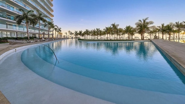 A large, curved swimming pool with a clear blue water surface, surrounded by palm trees and a hotel building with balconies. - Stunning Oceanview Two Story Condo - Beach - Gym (Nuevo Nayarit)