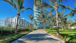 A pathway lined with palm trees leading to a modern hotel with a curved facade and multiple balconies. - Stunning Oceanview Two Story Condo - Beach - Gym (Nuevo Nayarit)
