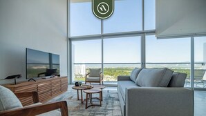 A modern living room with a large TV, comfortable seating, a coffee table, and a view of the cityscape through large windows. - Stunning Oceanview Two Story Condo - Beach - Gym (Nuevo Nayarit)