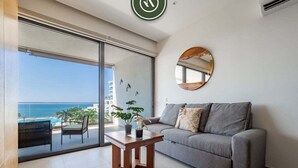 A modern living room with a grey sofa, a wooden coffee table, and a large window overlooking a balcony with a view of the sea. - Oceanview 1 BR Condo - Private Beach - Pools (Nuevo Nayarit)