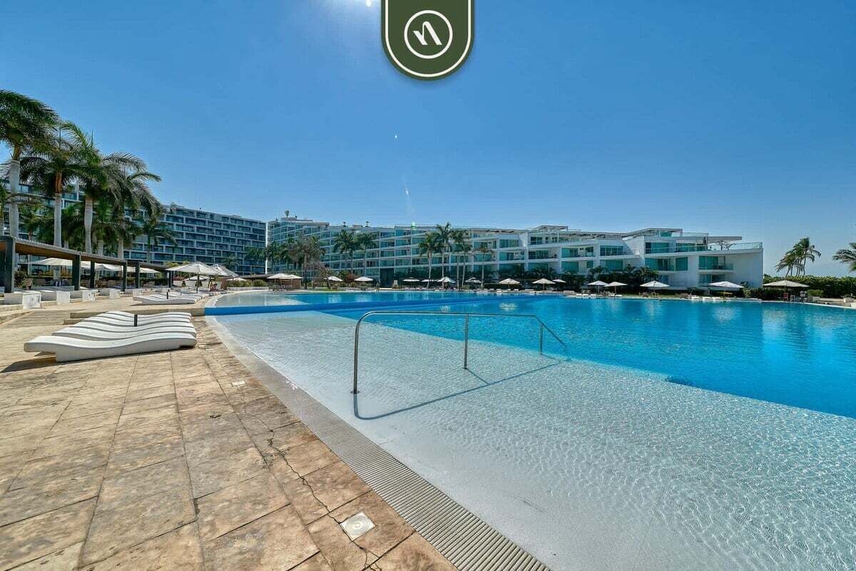 Una piscina con sillas de descanso, palmeras y un hotel al fondo.