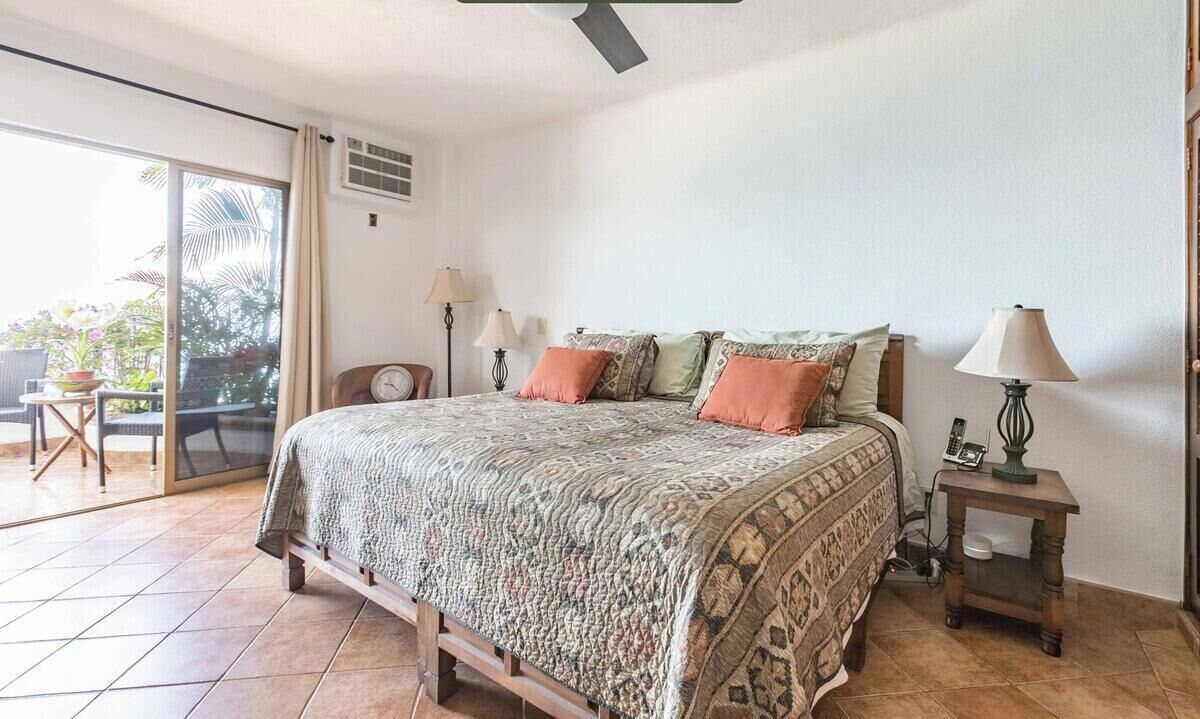 A bedroom with a bed, two bedside lamps, a small table, and a sliding glass door leading to an outdoor area with chairs and plants.