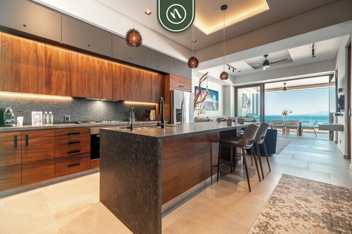Une cuisine moderne dotée d’un îlot central, de meubles en bois, de luminaires suspendus et offrant une vue sur l’extérieur.