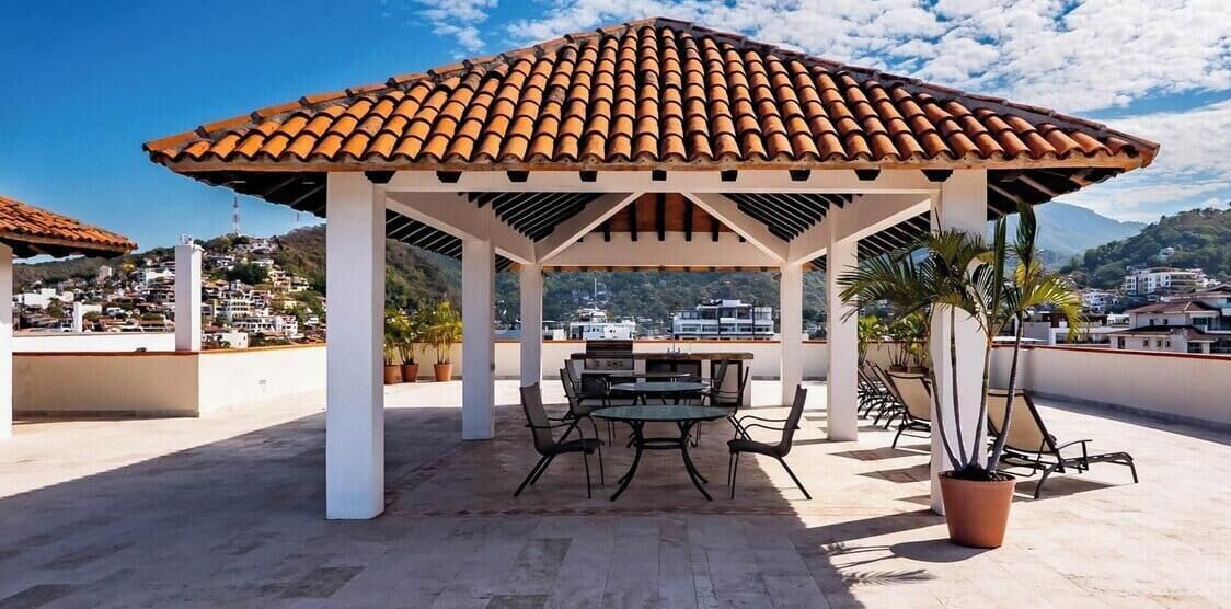 A covered outdoor seating area with a tiled roof, a table, and chairs, surrounded by potted plants.