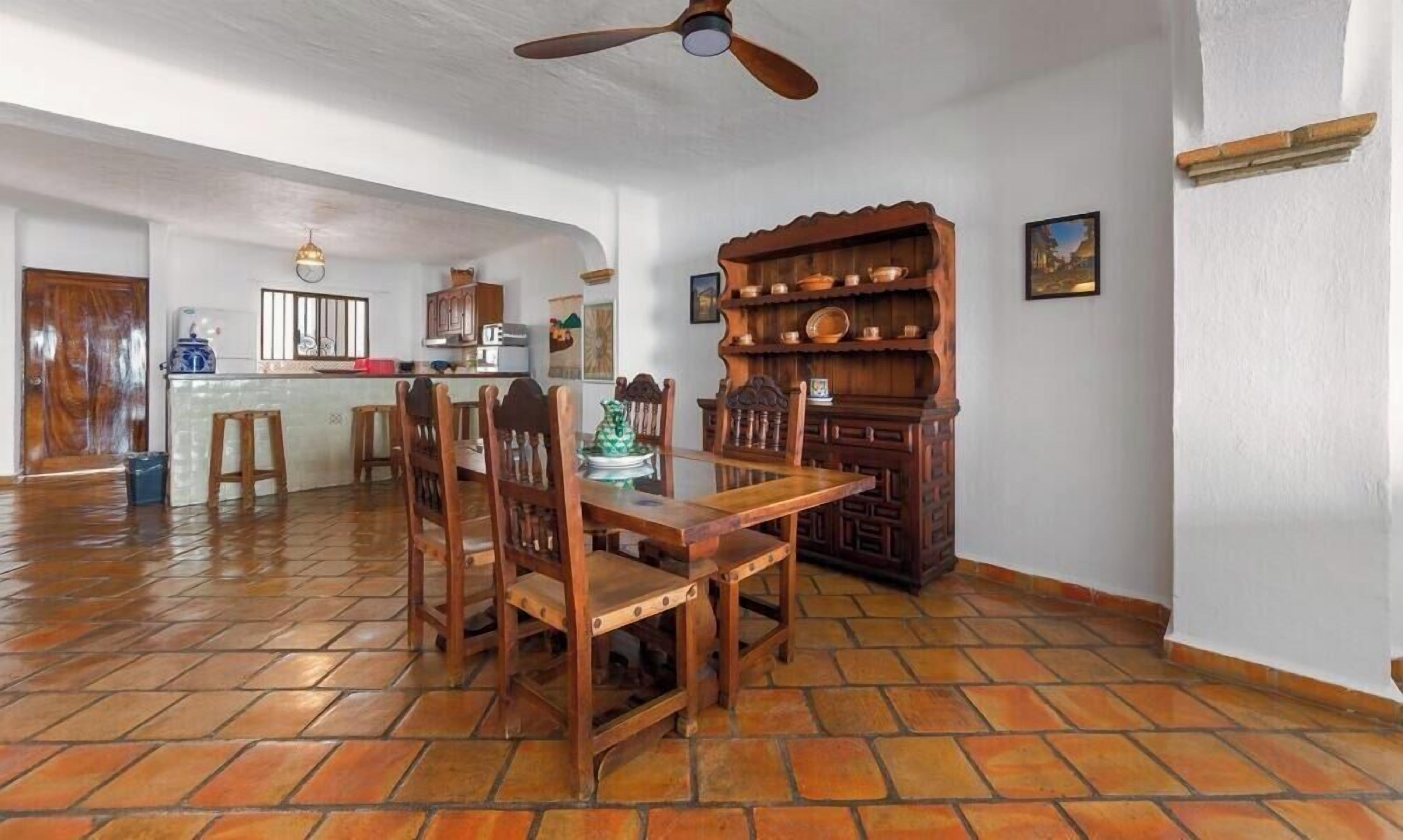A dining area with a wooden table and chairs, a kitchen in the background, and a ceiling fan.
