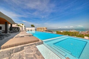 A swimming pool with a view of the ocean and cityscape, lounge chairs, and a covered seating area.
