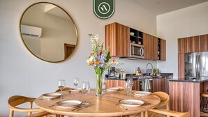 Ein Essbereich mit einem runden Holztisch, gedeckt für sechs Personen, umgeben von Holzstühlen. Ein Spiegel hängt an der Wand, und auf dem Tisch steht eine Vase mit Blumen.
