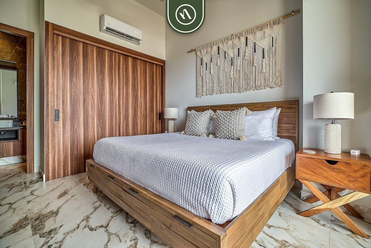 A bedroom with a wooden bed, a macrame wall hanging, and a nightstand with a lamp.